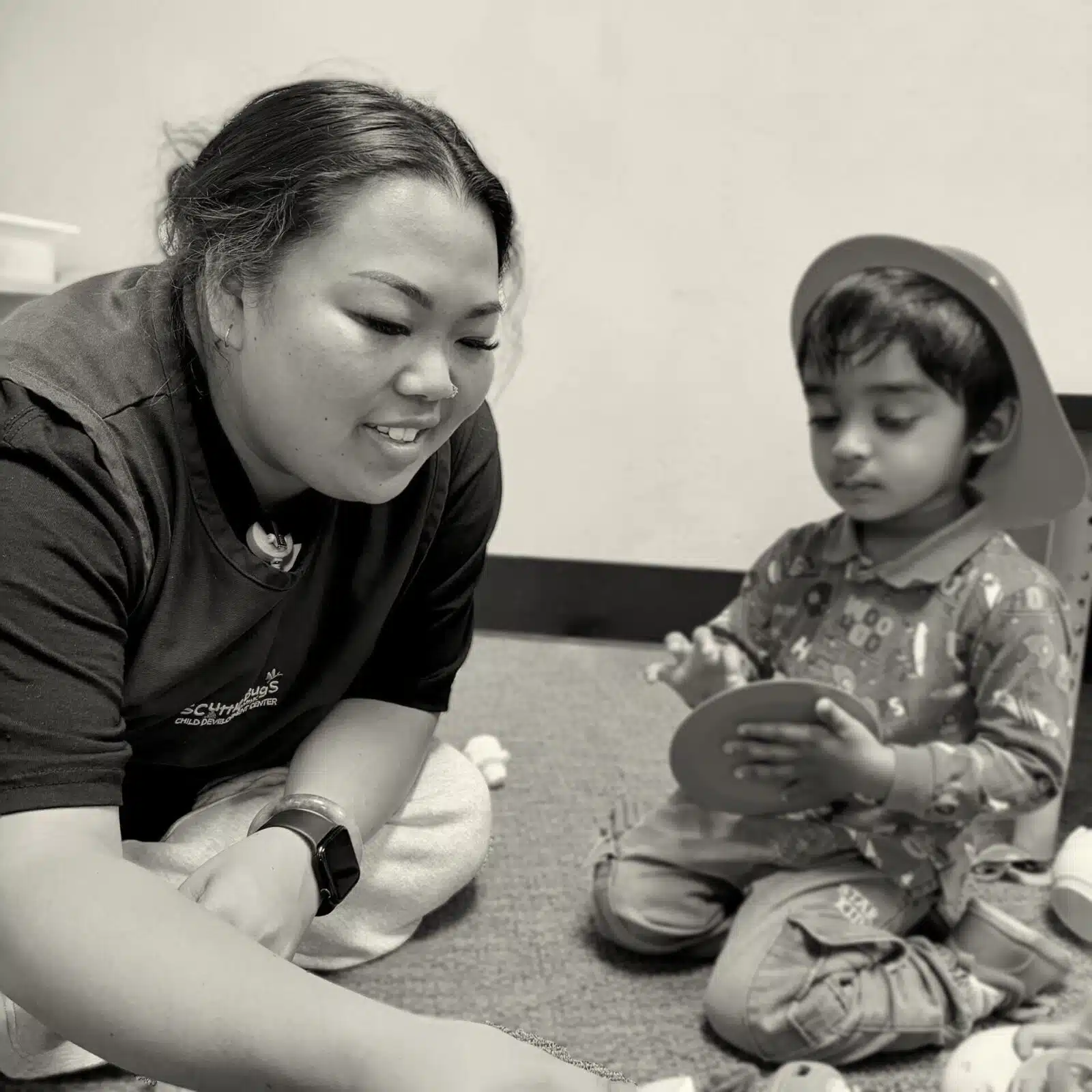 Caregiver and young child sit on a carpeted floor, focused on a round toy together.