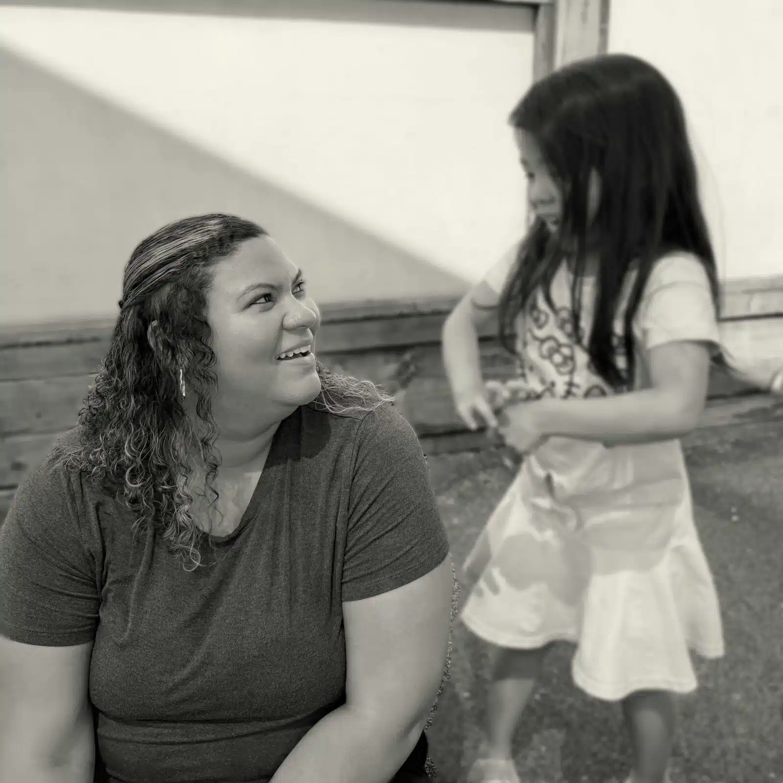 Foreground woman with curly hair smiling, wearing a dark shirt, in focus; a second girl in a light dress stands blurred in the background outdoors.