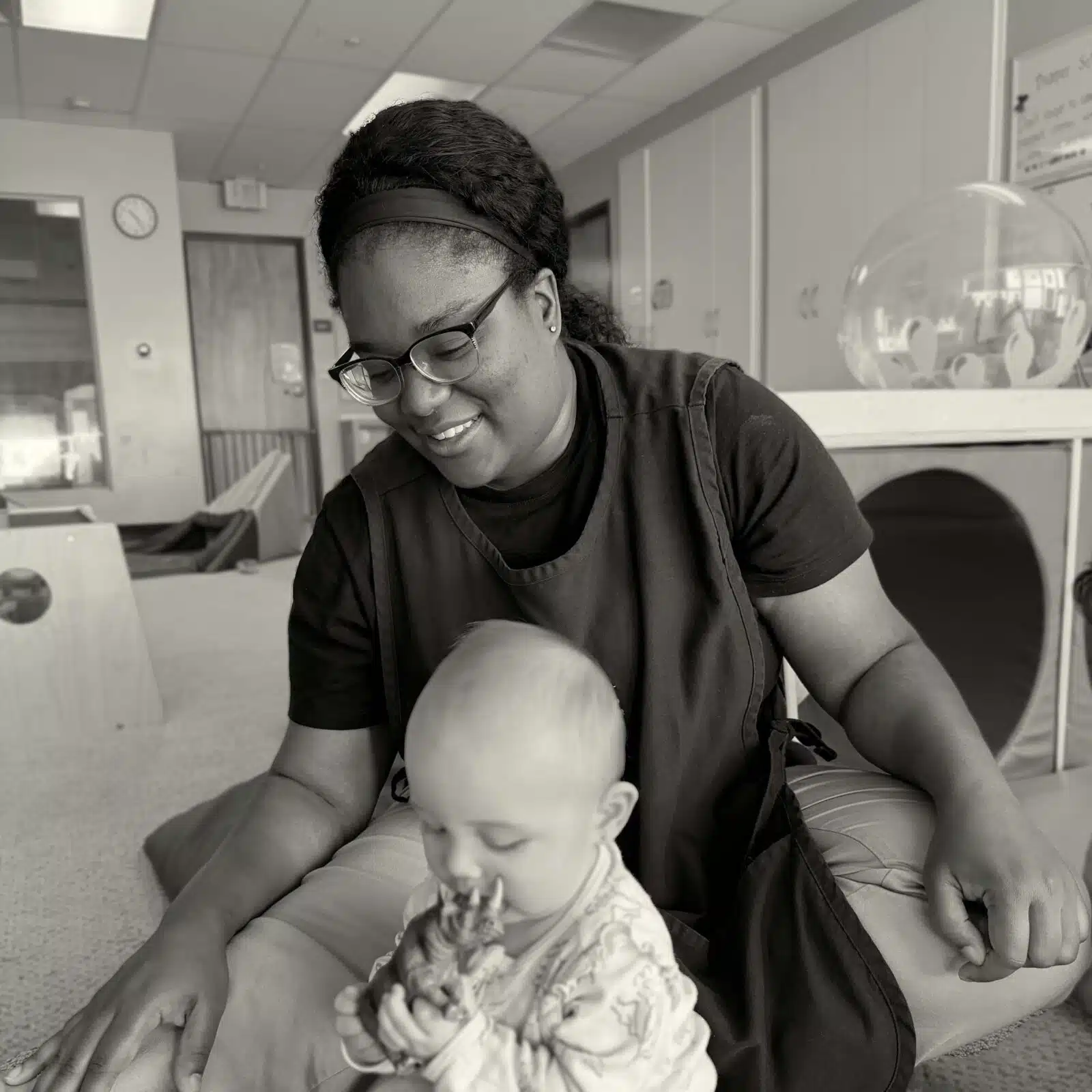 Healthcare worker in scrubs and glasses smiles while cradling a baby on her lap in a clinic.