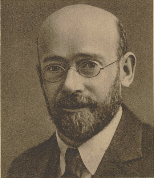 Sepia-toned portrait of a middle-aged man with round glasses, beard, and a suit looking at the camera.