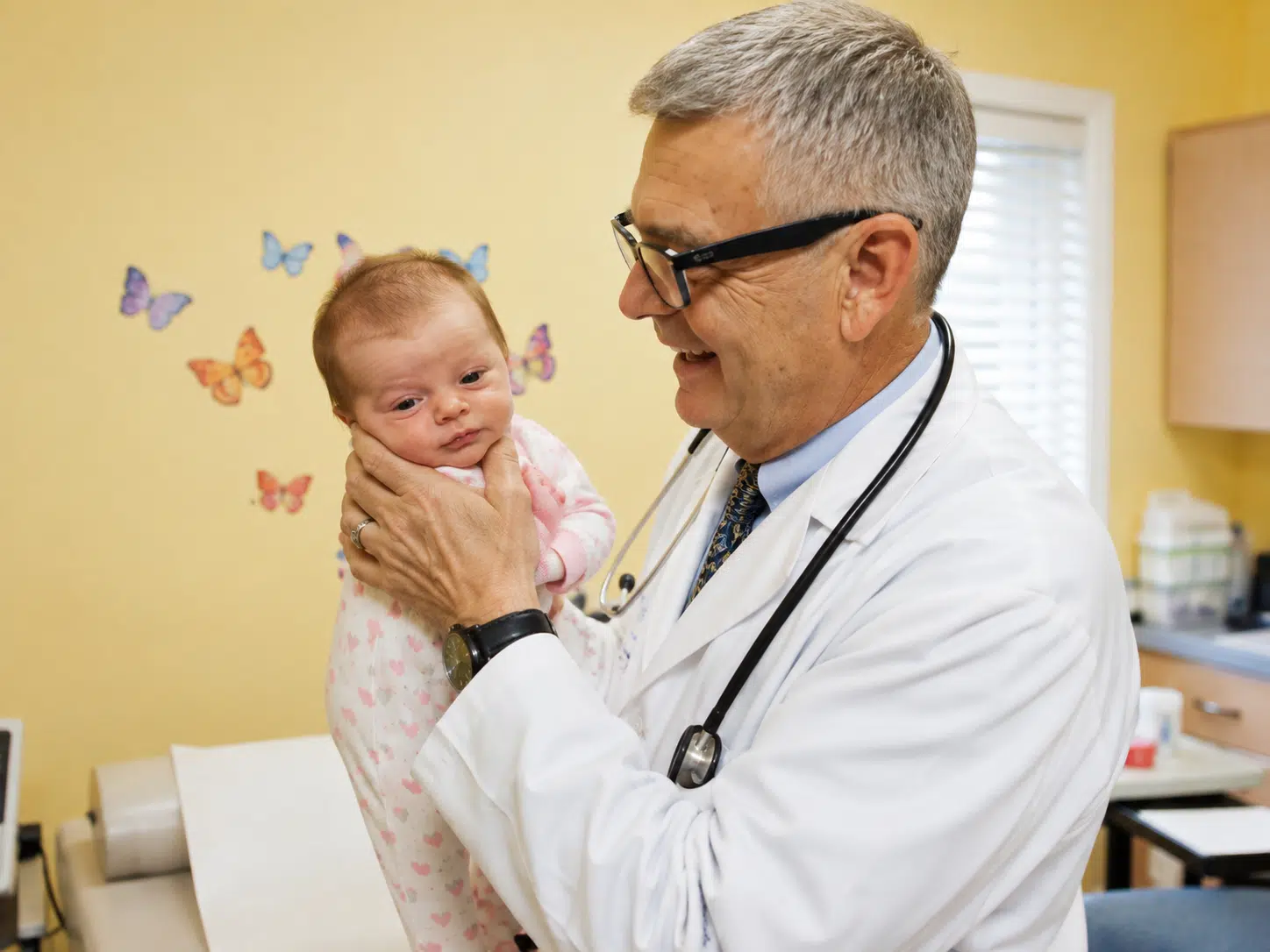 Pediatrician in a white coat holds a newborn baby and smiles in a bright clinic room.