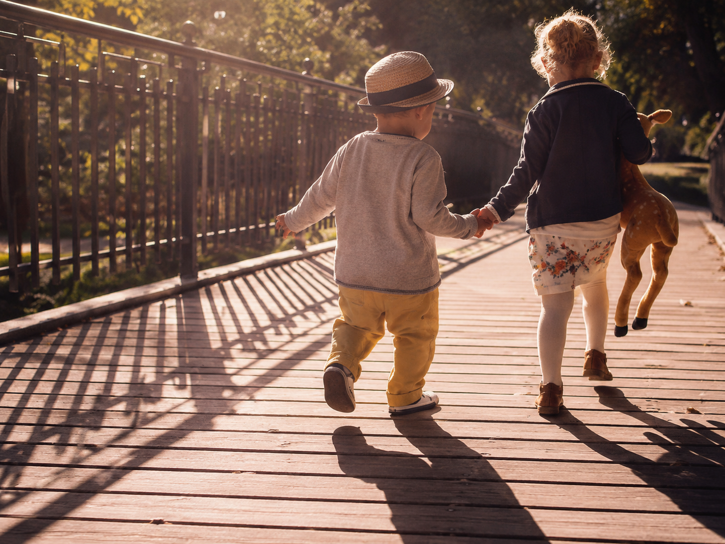 Two young children walk hand in hand on a sunlit wooden bridge, one holding a stuffed horse toy behind them.