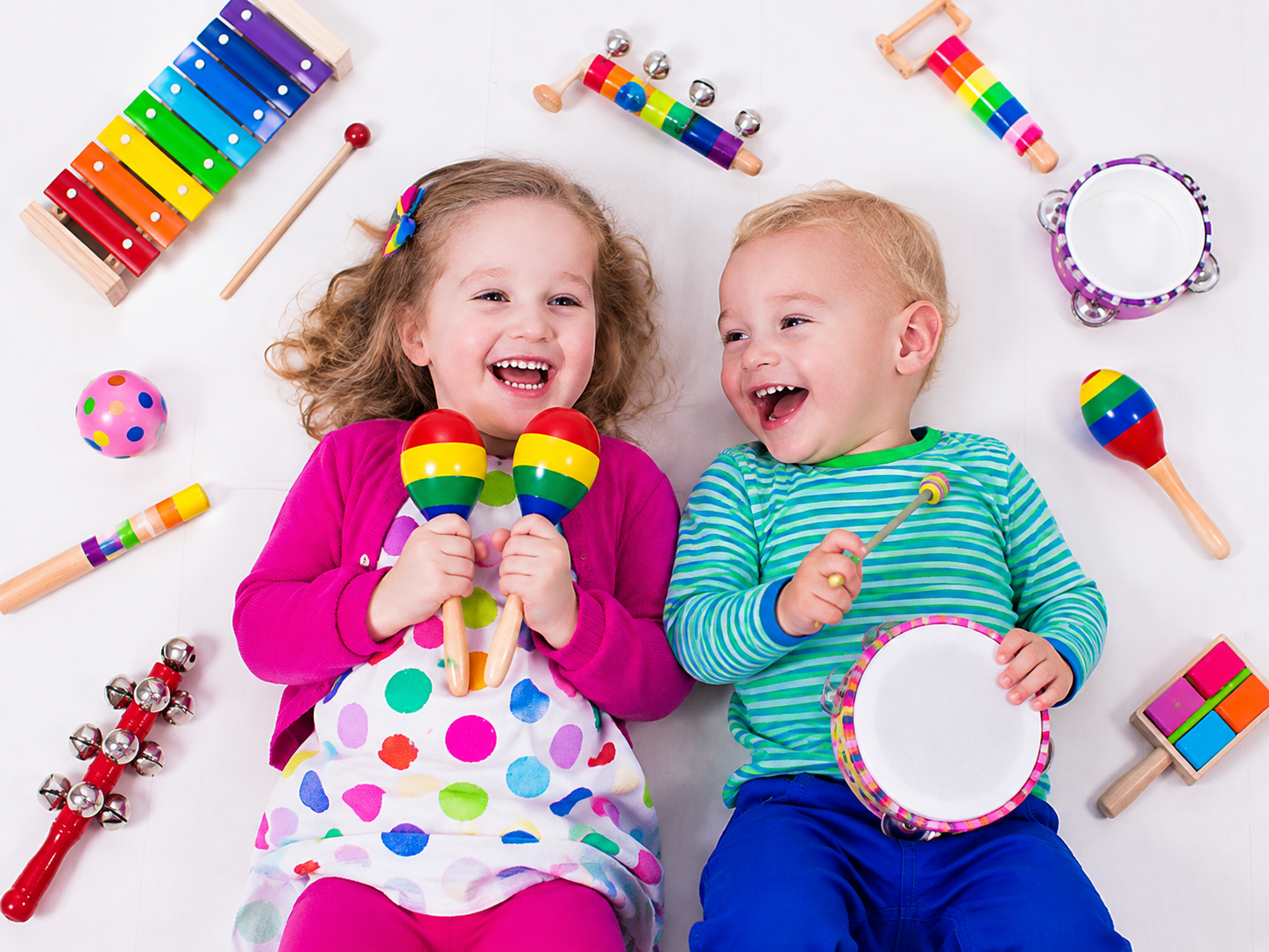 Two laughing kids lying on a white floor, surrounded by colorful musical toys, each holding rainbow maracas.