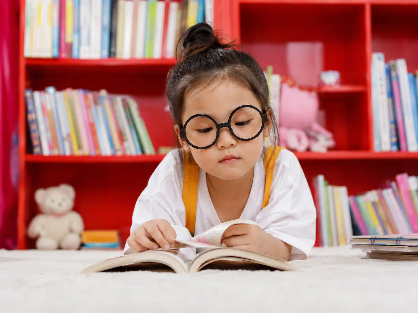 Young girl with round black glasses lying on a carpet, reading a book in a colorful library with red shelves and a teddy bear in the background