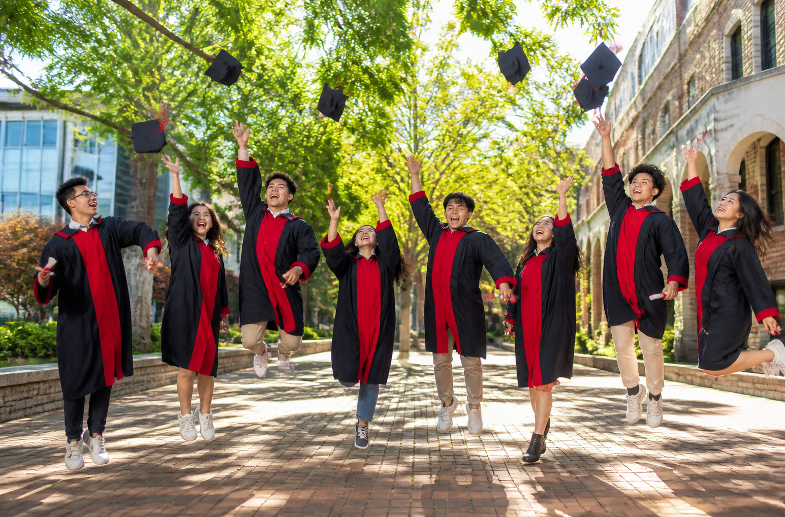 Group of graduates in black gowns with red trim jumping and cheering on a sunny campus path.