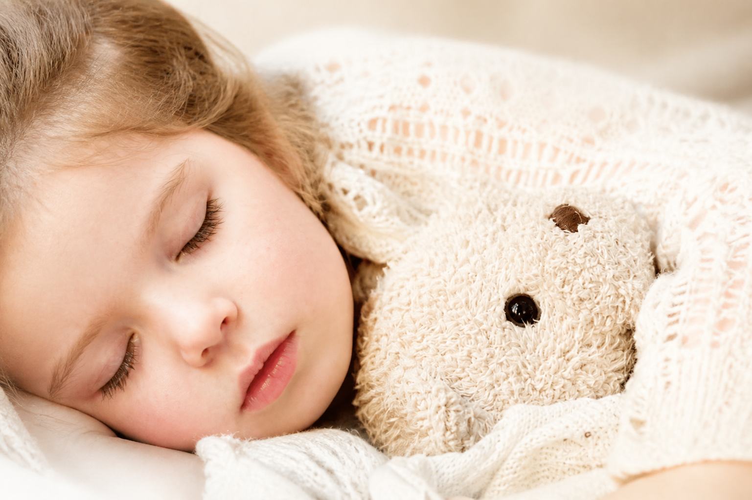 Close-up of a sleeping child cuddling a beige teddy bear under a cream blanket.