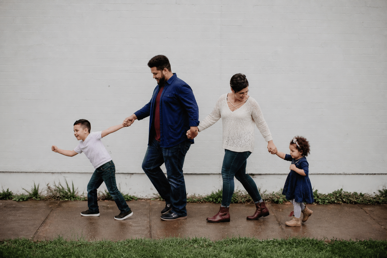 A father, mother, and two children hold hands and walk along a sidewalk in front of a pale gray wall, smiling as they go down the line.